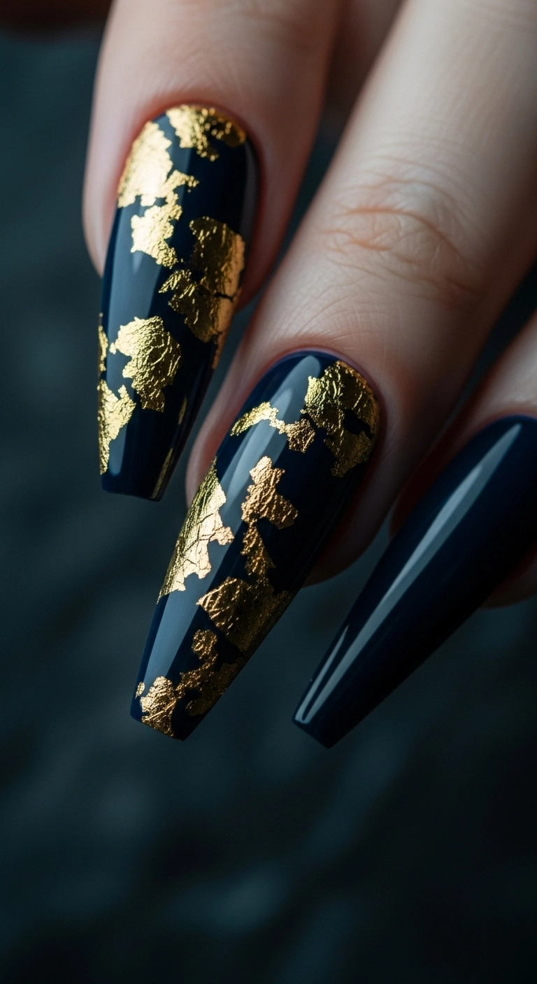 A close-up of a dramatic stiletto acrylic nail with gold foil accents on a dark navy blue base.