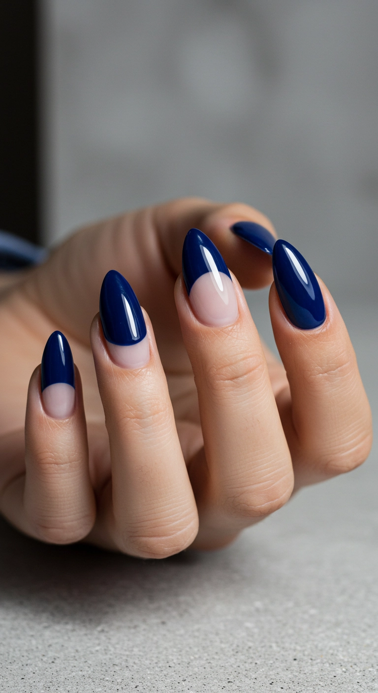 A close-up of almond nails with a deep navy blue polish, featuring a bare negative space half-moon at the cuticle.